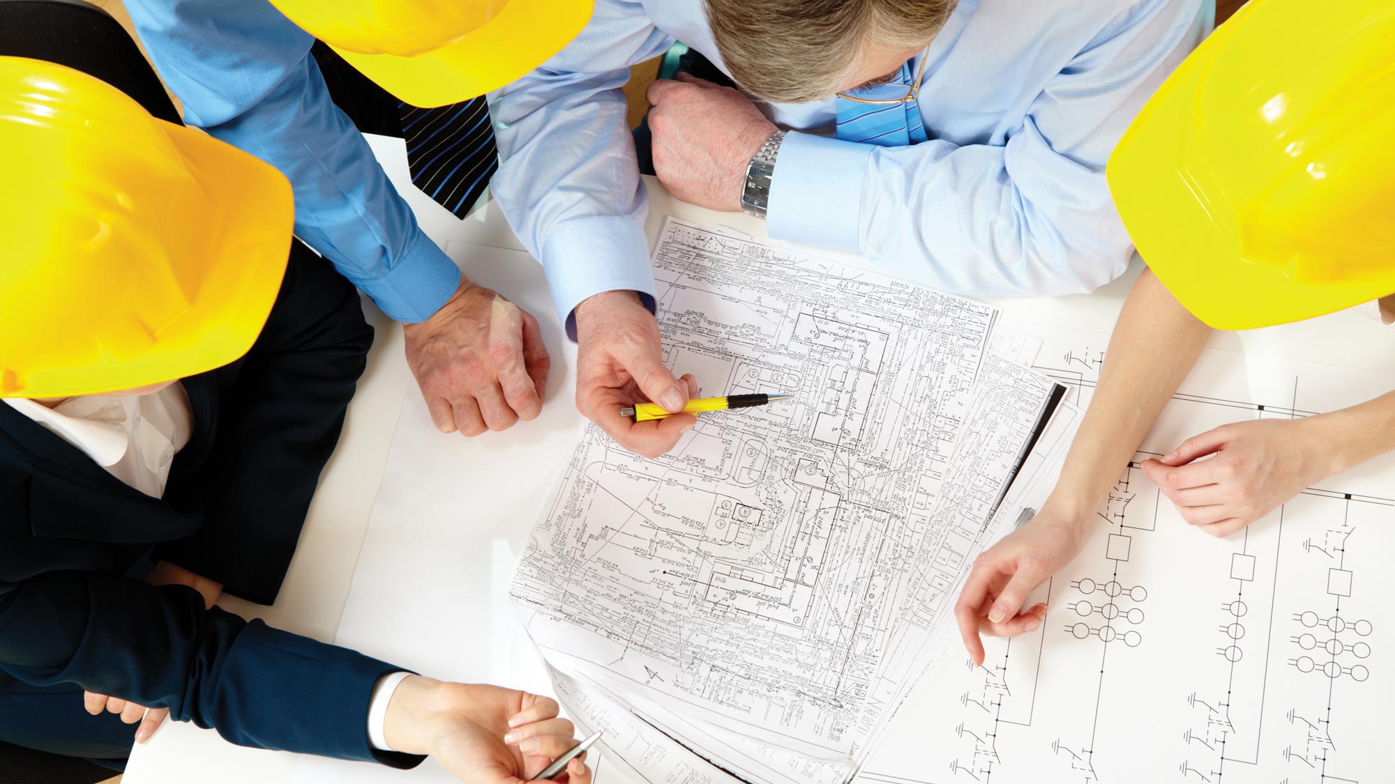A group of professionals in yellow helmets collaborating over detailed blueprints.