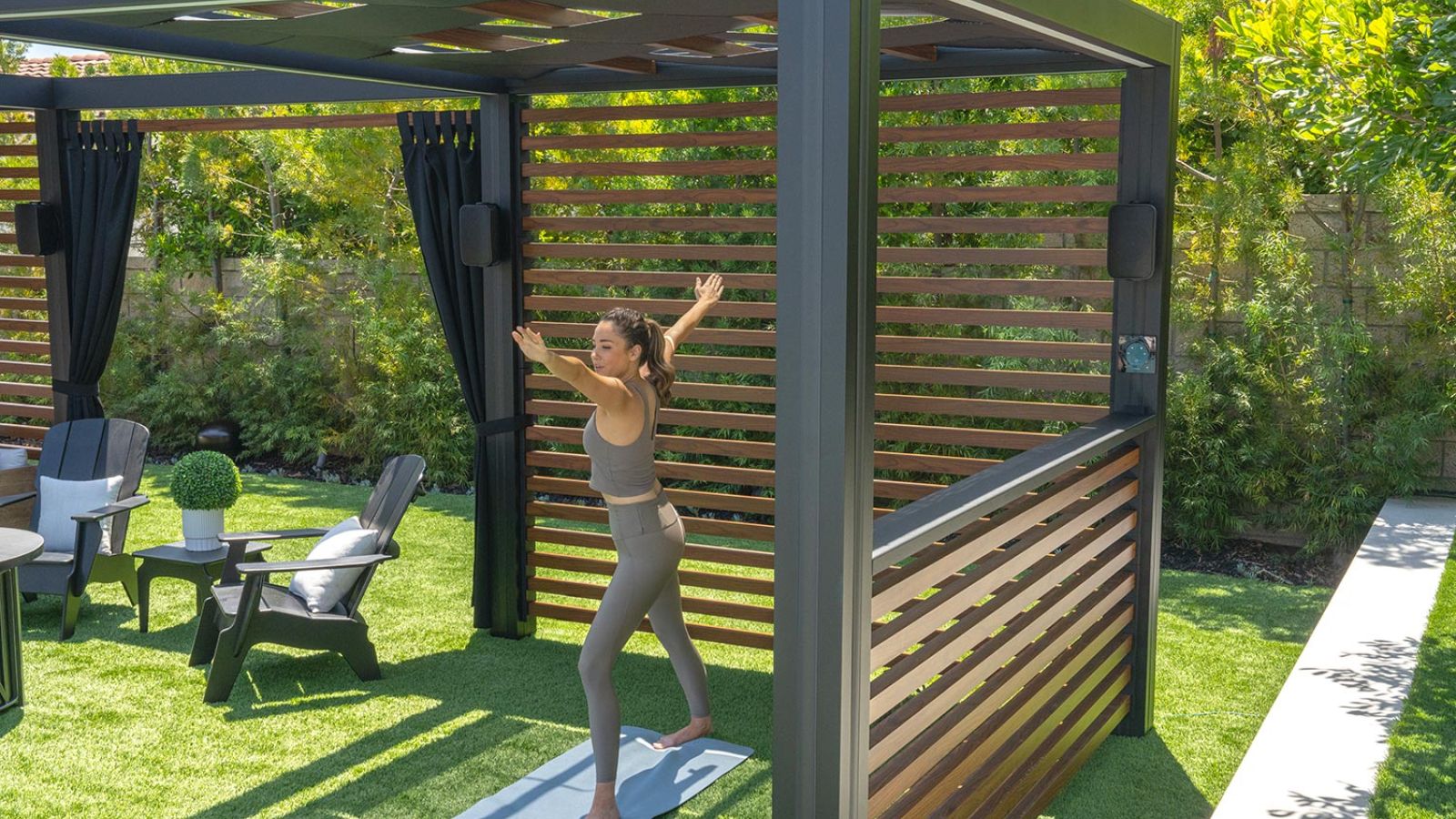 A group of people practicing yoga outdoors on a patio, following along with a fitness video playing on a mounted TV above an outdoor fireplace, surrounded by greenery.